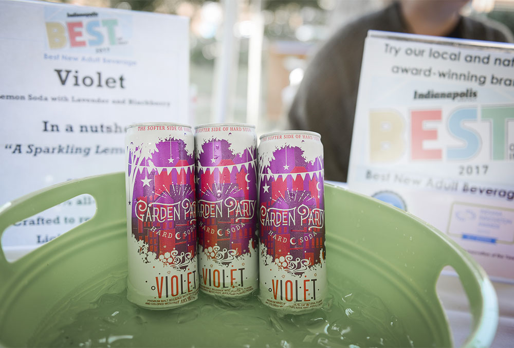 Photograph of Garden Party Botanical Hard Soda cans, bathing in a pool of ice water in a light green bucket. Featured in the background are two posters showing awards such as Indianapolis's best new adult beverage of 2017.