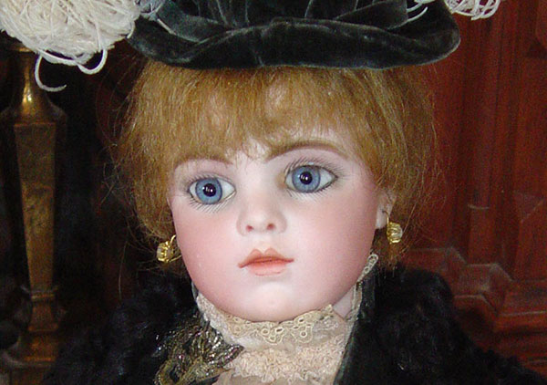 This close up photograph of the doll shows some finer details of the face, hat, and dress. Doll has Blonde hair and blue glass eyes. Her head shoulders, and forearm are bisque with upper arms & body thighs are kidskin leather. The lower legs and feet are celluloid. Tiffany mounted, fake pearl pierced earrings hand from her pierced ears. She is wearing a late nineteenth century style black dress with a feathered hat.