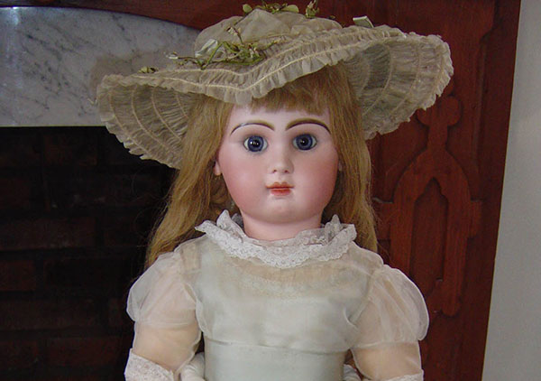 Photo of Baby Mckee's jumeau doll. The head is bisque and composition body has jointed arms and legs, blue glass eyes and long curled blond hair. The doll has pierced ears with imitation ruby earrings.