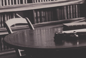 Photo of a desk in an office, darkly toned, in front of a bookcase lined with large books. On the desk are two thick books, next to each other.