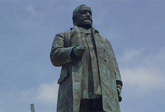Image of Benjamin Harrison Statue, made of copper and turned green from age. Harrison wearing a long coat and holds his arm by his side at a ninety degree angle at chest level. The sky behind his is pale blue with thin clouds.