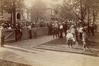 Photograph of crowd in front of Harrison's North Delaware Street home in 1888. Wider view showing crowd in front of Delaware Street home and showing the houses to the north. Brick driveway and sidewalk in foreground.