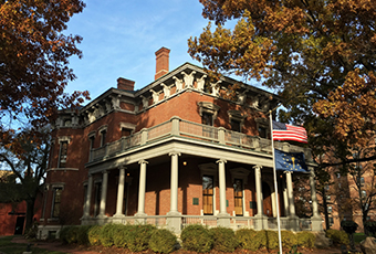 Image of presidential mansion during fall at dusk. The house is slightly obscured and shaded by surrounding oak trees. The house is made of brick with white accents, three floors in height. A flagpole flying the american flag stands out front.