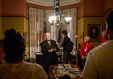 photograph of an actor playing benjamin harrison, conferring with a tour group in his office space.