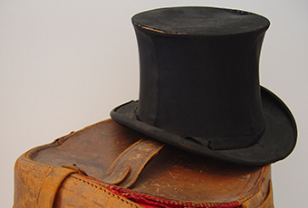 Benjamin Harrison's silk top hat on top of a leather carrying case. Black satin or silk hat. the hat is 6 inches tall with a slight bowed flare toward the middle the stack; seams on both sides; a band to cover the joining of stack and brim is missing except on the right front; a partial bow is placed on the right side; stitches are showing where band would have been.