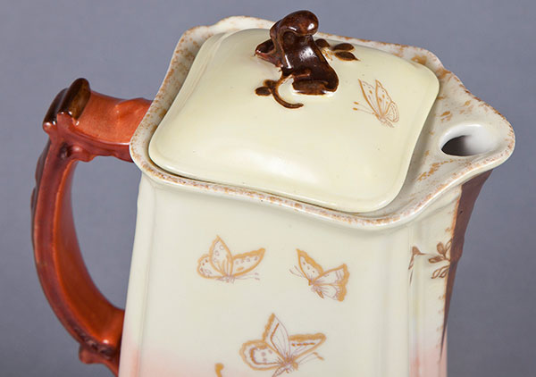 close up photo of the top of the chocolate pot. The handle is painted brown with stems and leaves protruding from it to emulate the natural aesthetic of the rest of the pot. The lip of the pot has gold trimming.