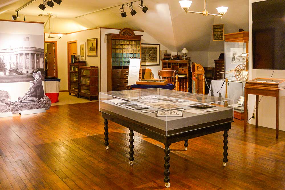 Photograph of the third level of the benjamin harrison home. Several display cases, paintings, and bookcases can be seen in the image. The floor is wooden and the ceiling is lower than the rest of the home.