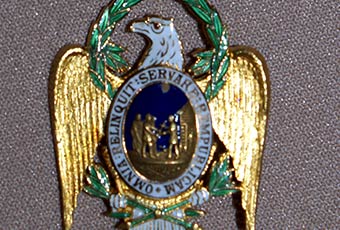 Photo of harrison's badge. bears a predominantly light blue ribbon, with white edging, holding an eagle with golden wings and wreath around head. The center is a picture of figures against a blue background, encircled by the text 