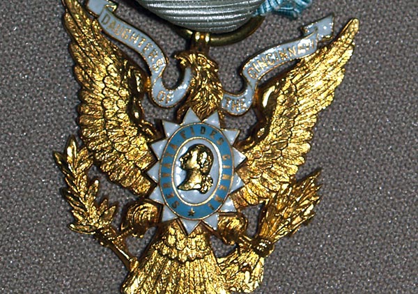 Daughter of the Cincinnati badge. Predominantly white ribbon with blue edging holding a golden eagle. An emblem in the center of the eagle contains a golden bust of a male figure and is encircled by the text 