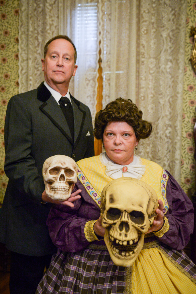 photo of two candlelight theater actors, one man standing on the left and one woman seated on the right. each is holding a human skull in their hands.