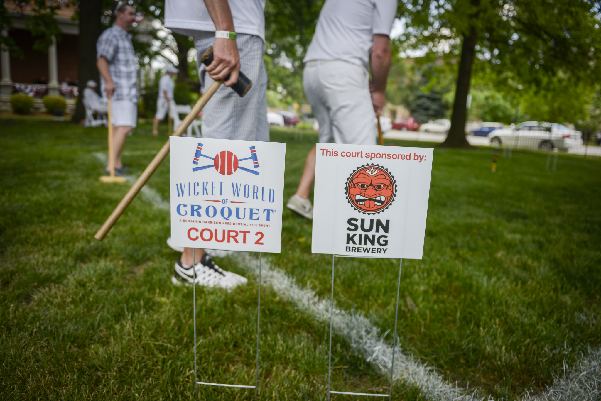 Photo of several men playing croquet. In the foreground, a man stands with his mallet in his hands while a man in front of him lines up a shot. A man in the background can be seen observing the game. In the very front are two signs, one sporting the wicket world of croquet logo, showing that this is court 2. the other sports the logo of sun king brewery, a sponsor of the benjamin harrison site.