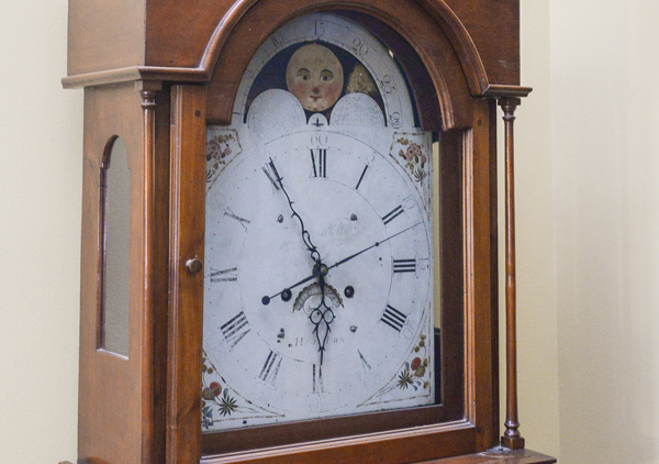 Tall case or grandfather clock made by George Woltz in Hagerstown, Maryland between 1800 and 1812. Clock has moon dial at top and a date dial below the center of the hands, and strikes on the hour. Cherry cabinet with front legs that curve out . There is a spindle on either side of the face. Rounded center at the top with rosette design. Top corners come up to pointed spindles. Clock face is a half circle at top with the dates above the moon dial, Roman numerals and painted floral designs in four corners, and glass door.