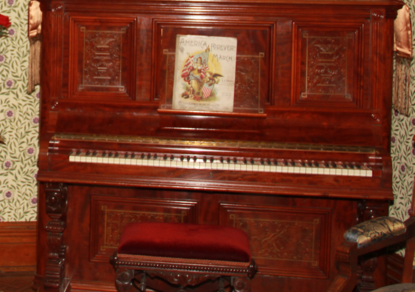 Wooden upright piano. There is a silver plaque mounted above the keyboard with manufacturer's name and information. The wood panels on each side of front have an incised design. Center panel has incised design of violin and mandolin crossed surrounded by flowers and leaves.
