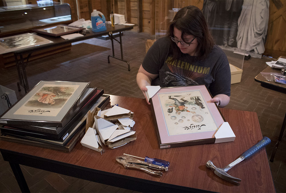 Photo of curator emily hannawalt. in the photo, she can be seen sitting at a desk with a hammer and other tools. she is framing several paintings using white corners, which are haphazardly lying on each other while a pile of paintings is neatly stacked nearby.