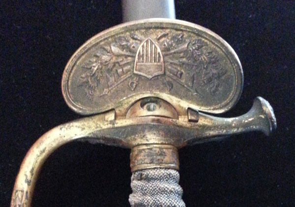Benjamin Harrison's Civil War sword. Handle or grip is covered with a leather or skin. The pommel at the top of the hilt is brass or gold color metal with a rosette or flower design on top and an eagle on one side. The grip has an extended handle of brass or gold color metal with design. The bottom of the hilt or rain guard is hinged on one side. The guard is brass or gold color metal with a shield design on the hinged side and an eagle design on the other side. There is a finger guard opposite the handle that extends out about 1 1/2