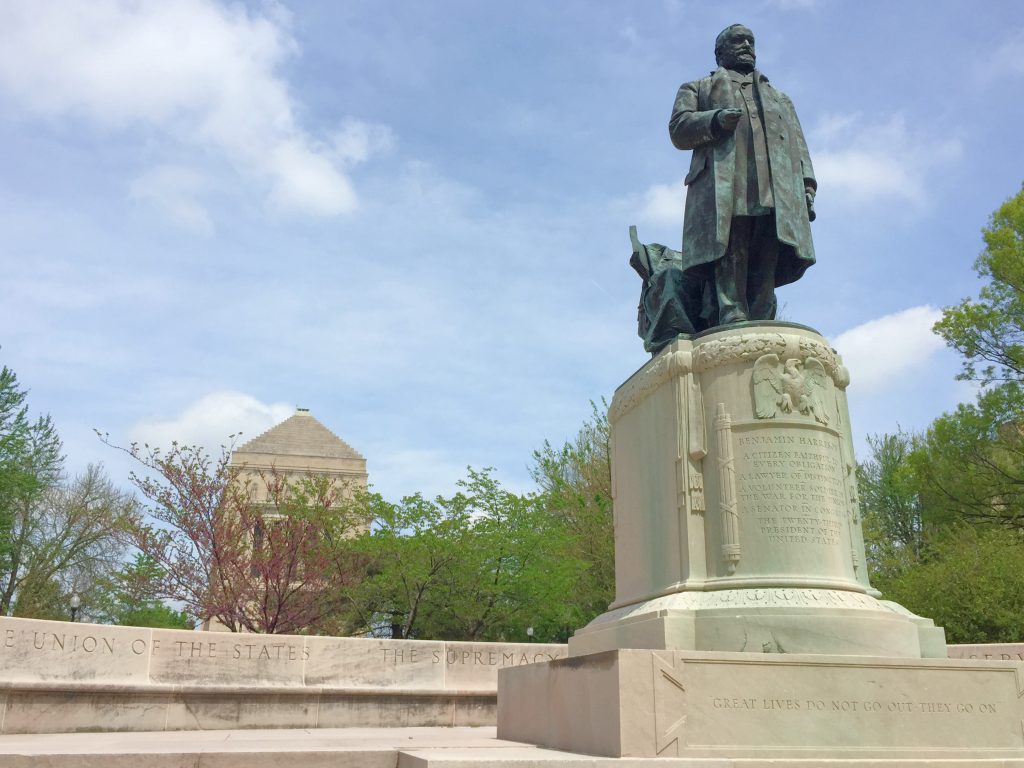 Photograph of Benjamin Harrison's statue, forged from copper and turned green over time. The sky is a pale blue and leafy green trees cascade against the background. The engraving at the base of the statue reads, "Great lives do not go out, they go on". A quote famously said by President Harrison.